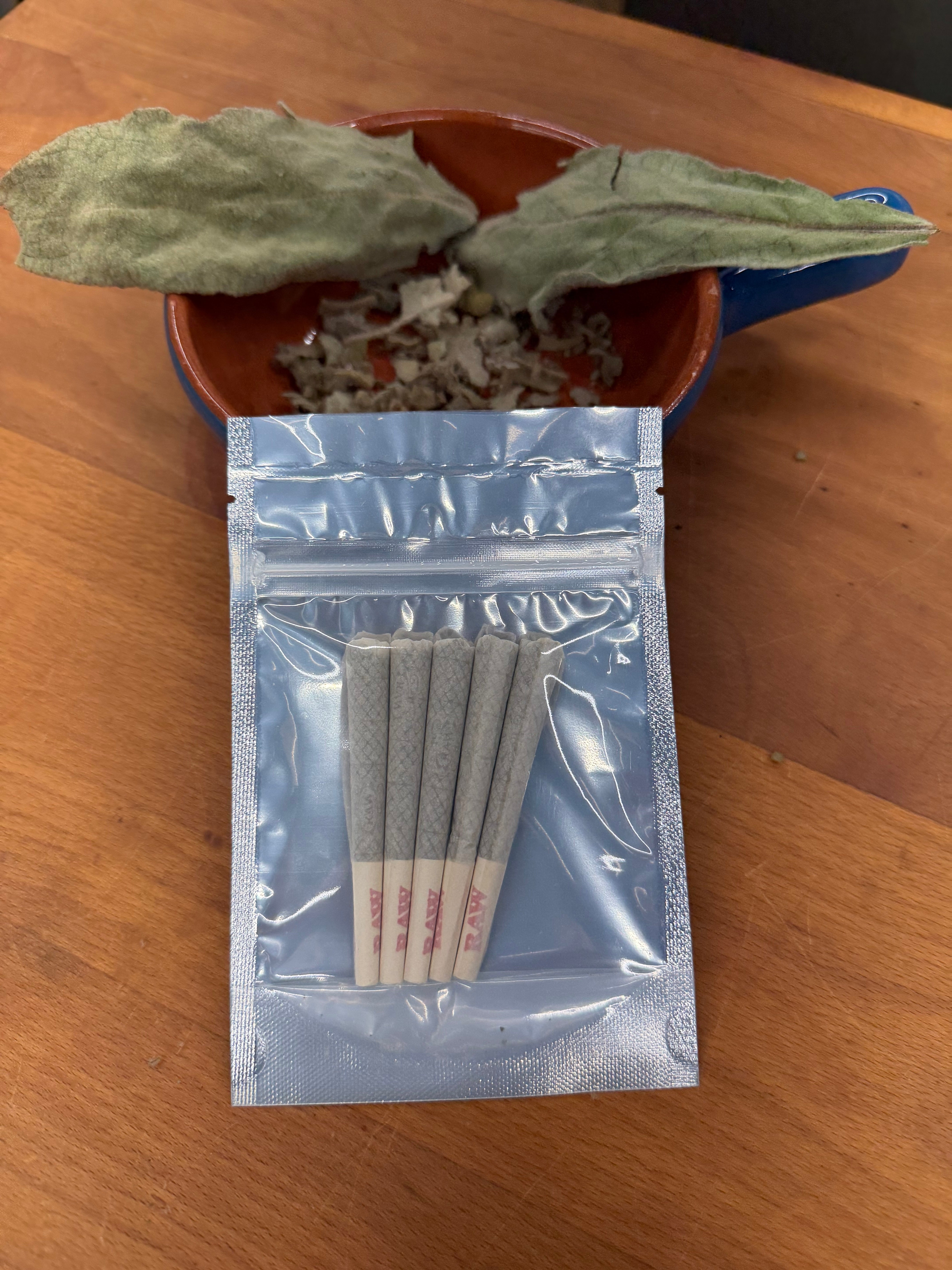 Two whole dried mullein leaves and crumbled leaf in a blue enamel dish above an open mylar bag holding five prerolls